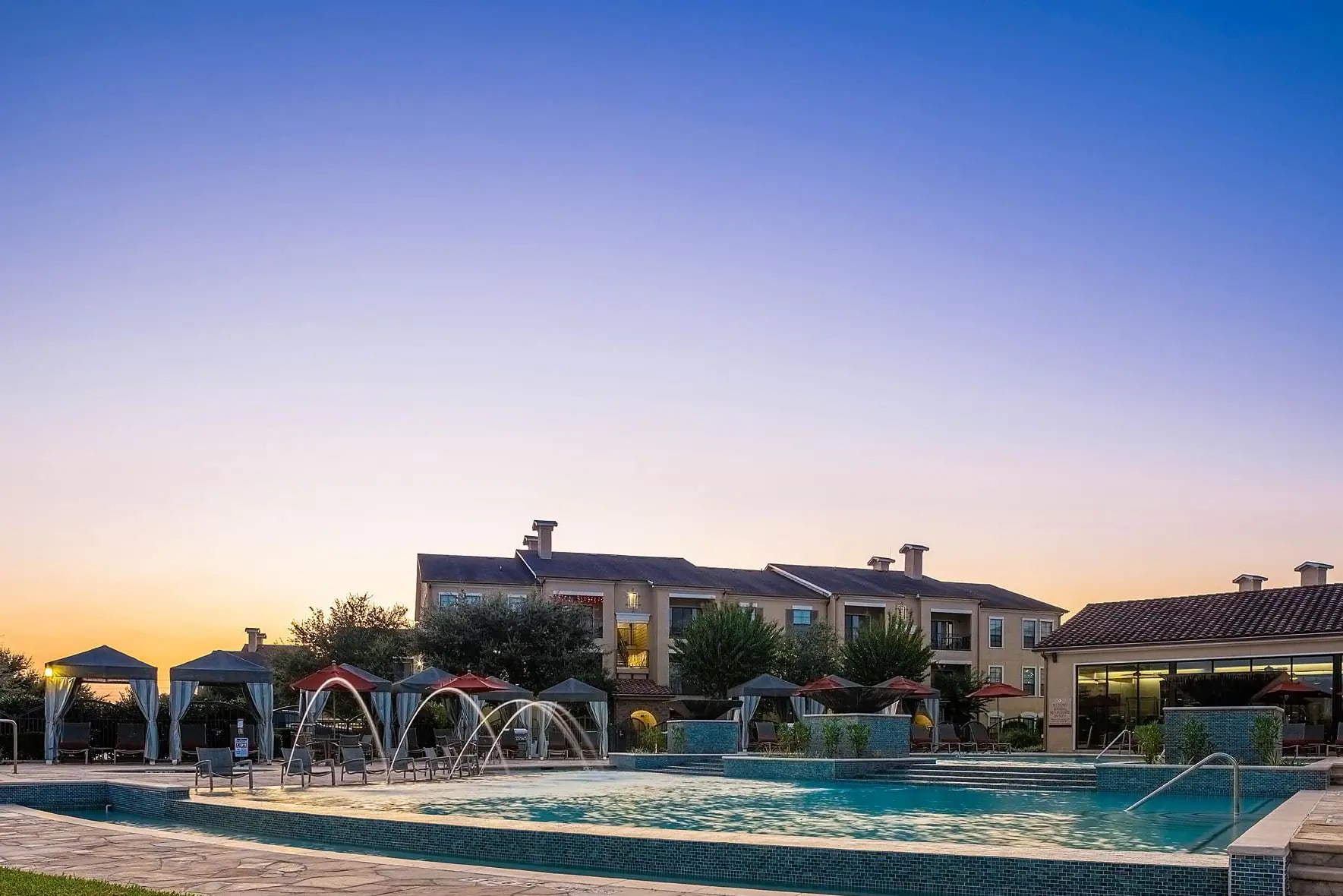 Resort pool with blue water, white cabanas, arch water features, and multi-story residential buildings at sunset.
