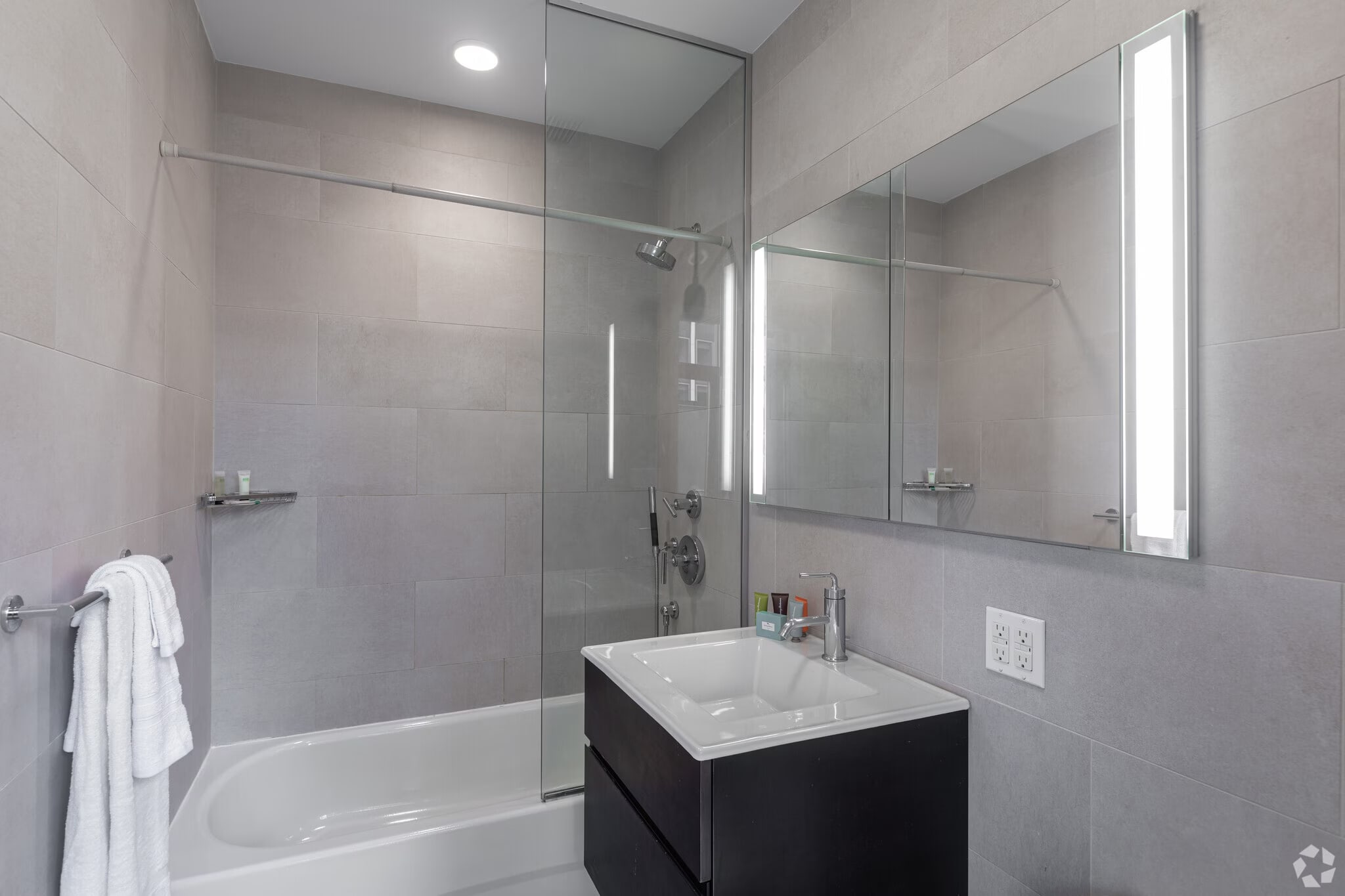 Contemporary bathroom with gray tile walls, glass-enclosed shower, white soaking tub, black floating vanity with white sink, and lighted mirror.