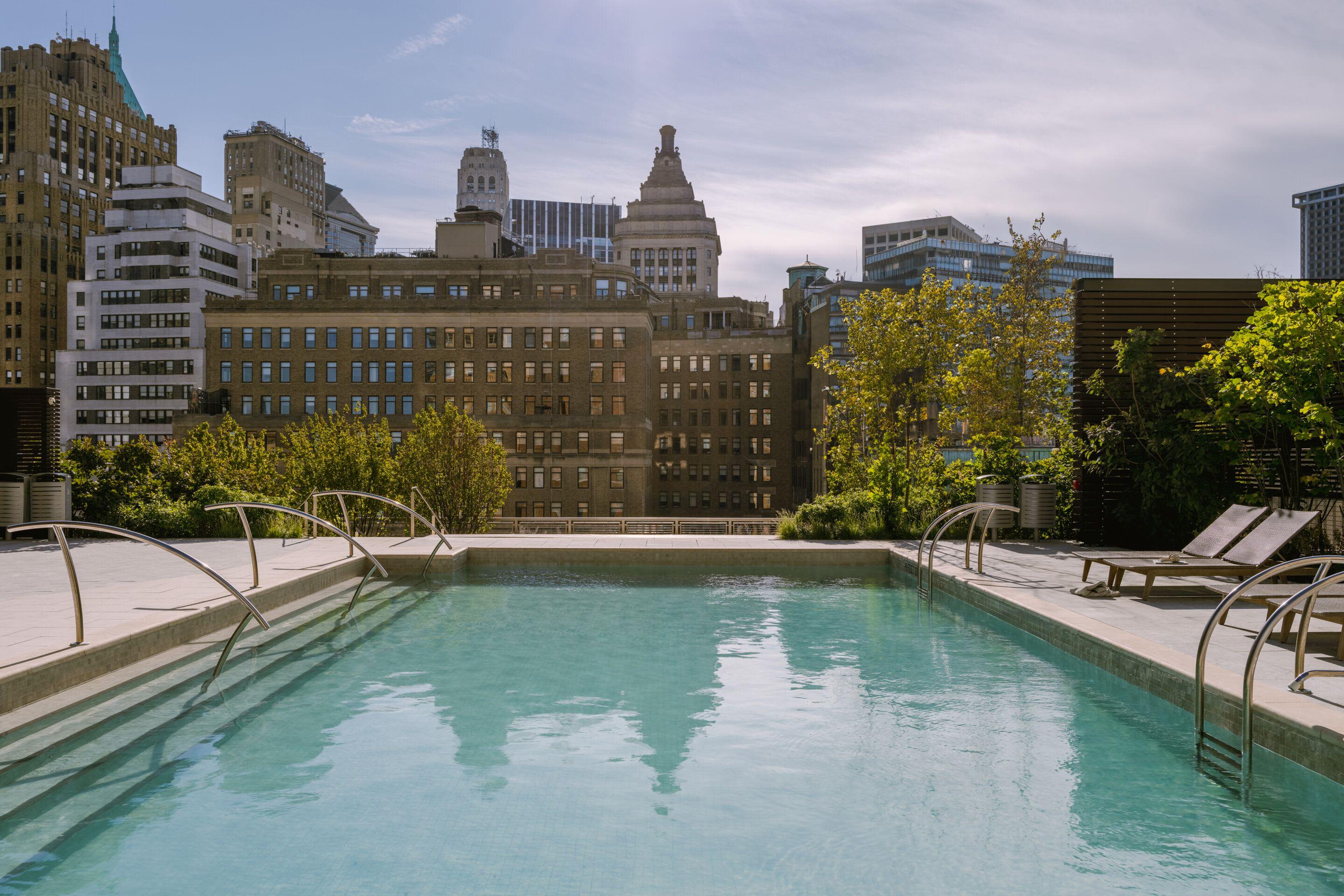 Rooftop swimming pool with turquoise water, lounge chairs, and metal railings, with downtown Manhattan skyline and historic buildings in background.