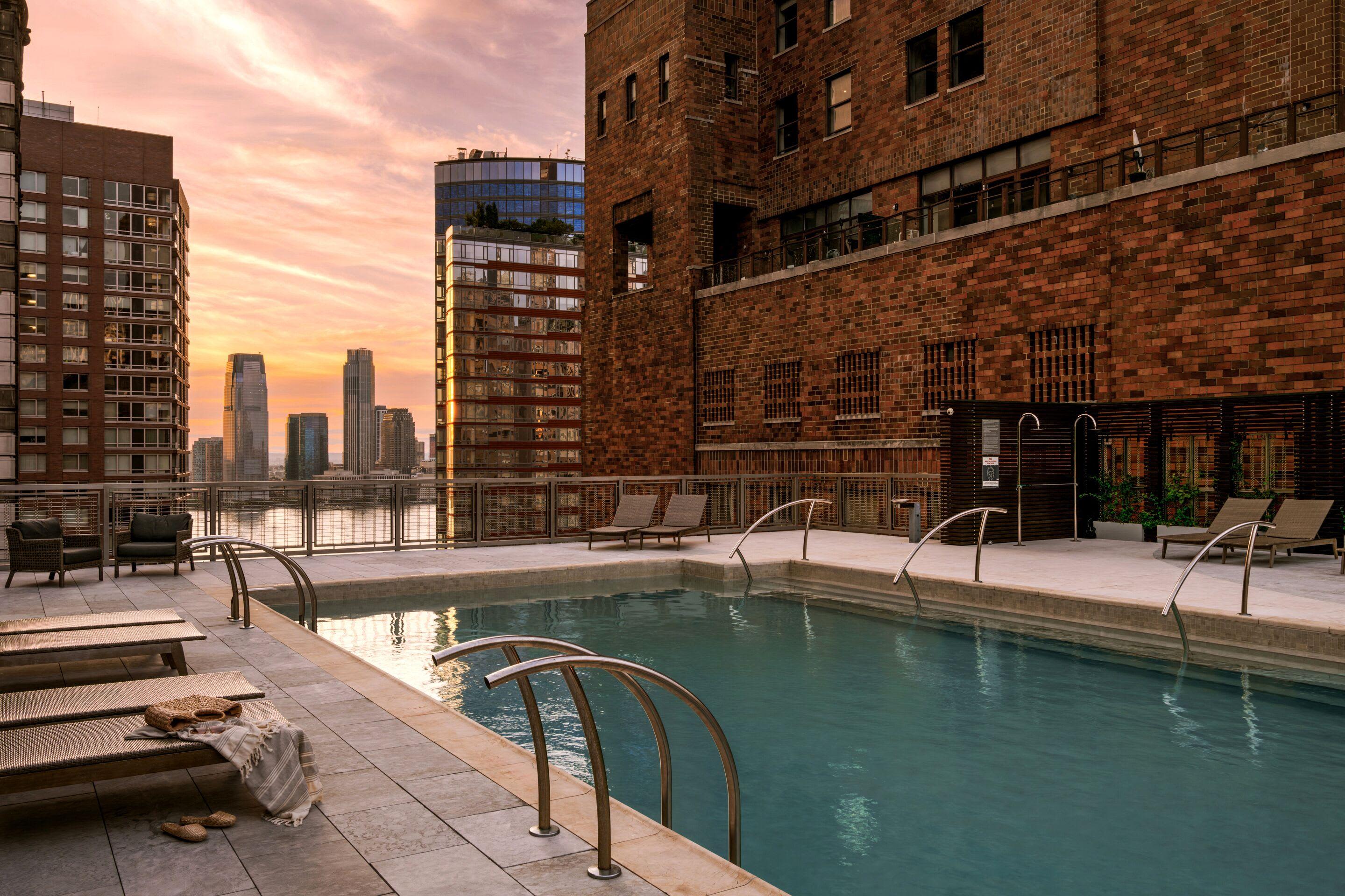 Rooftop pool at sunset with lounge chairs, metal railings, brick building facade, and Manhattan skyline in background.