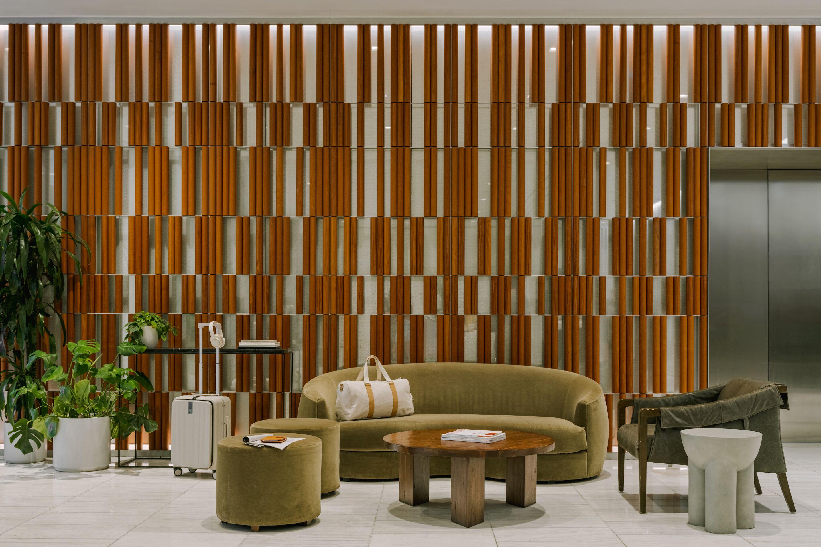 Curved olive-green sofa with round wood coffee table in modern lobby, wood-slat wall backdrop, potted plants, and metal elevator doors.