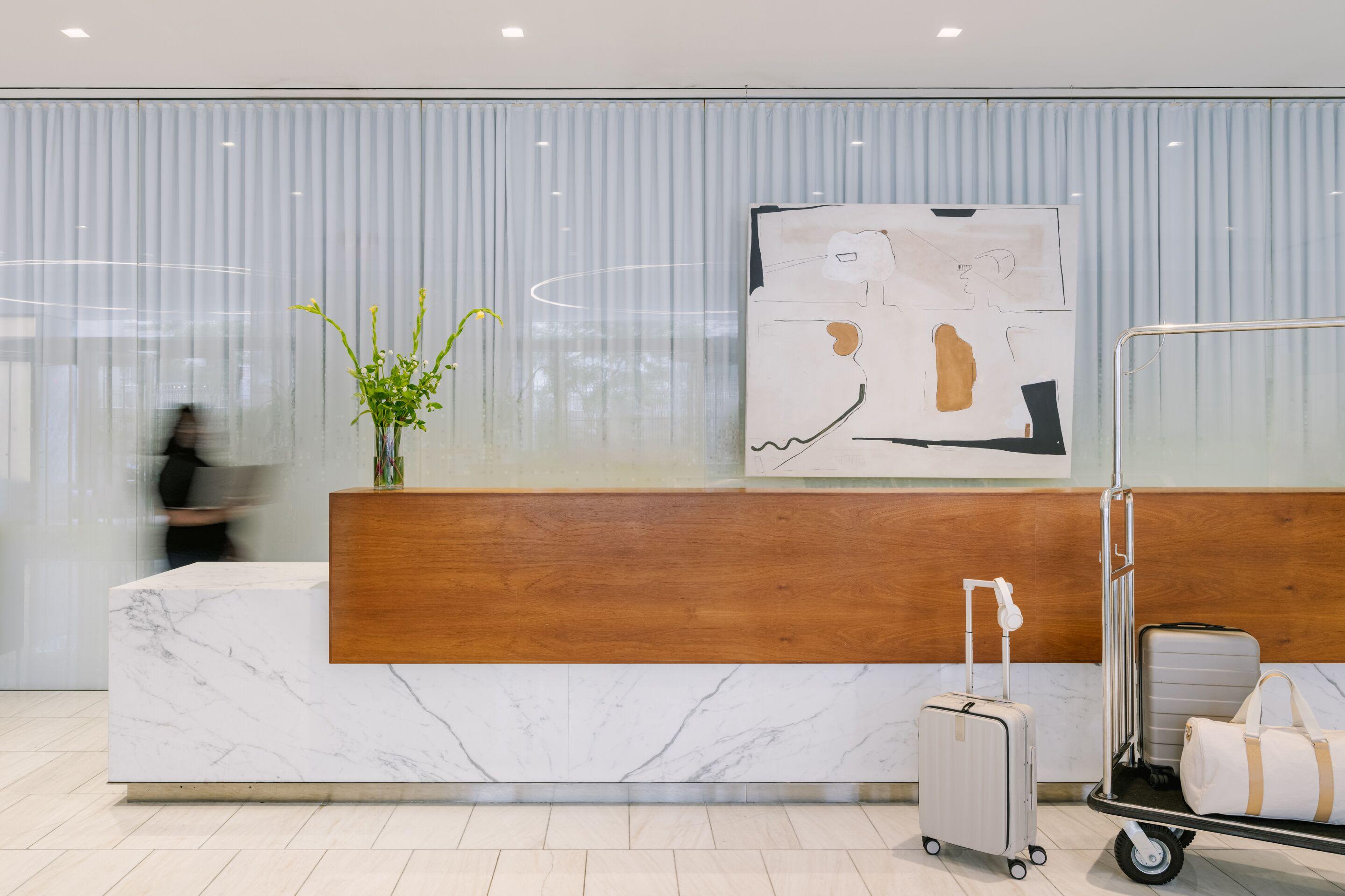 Hotel lobby with marble and wood reception desk, fresh flowers in glass vase, abstract wall art, and guest luggage.
