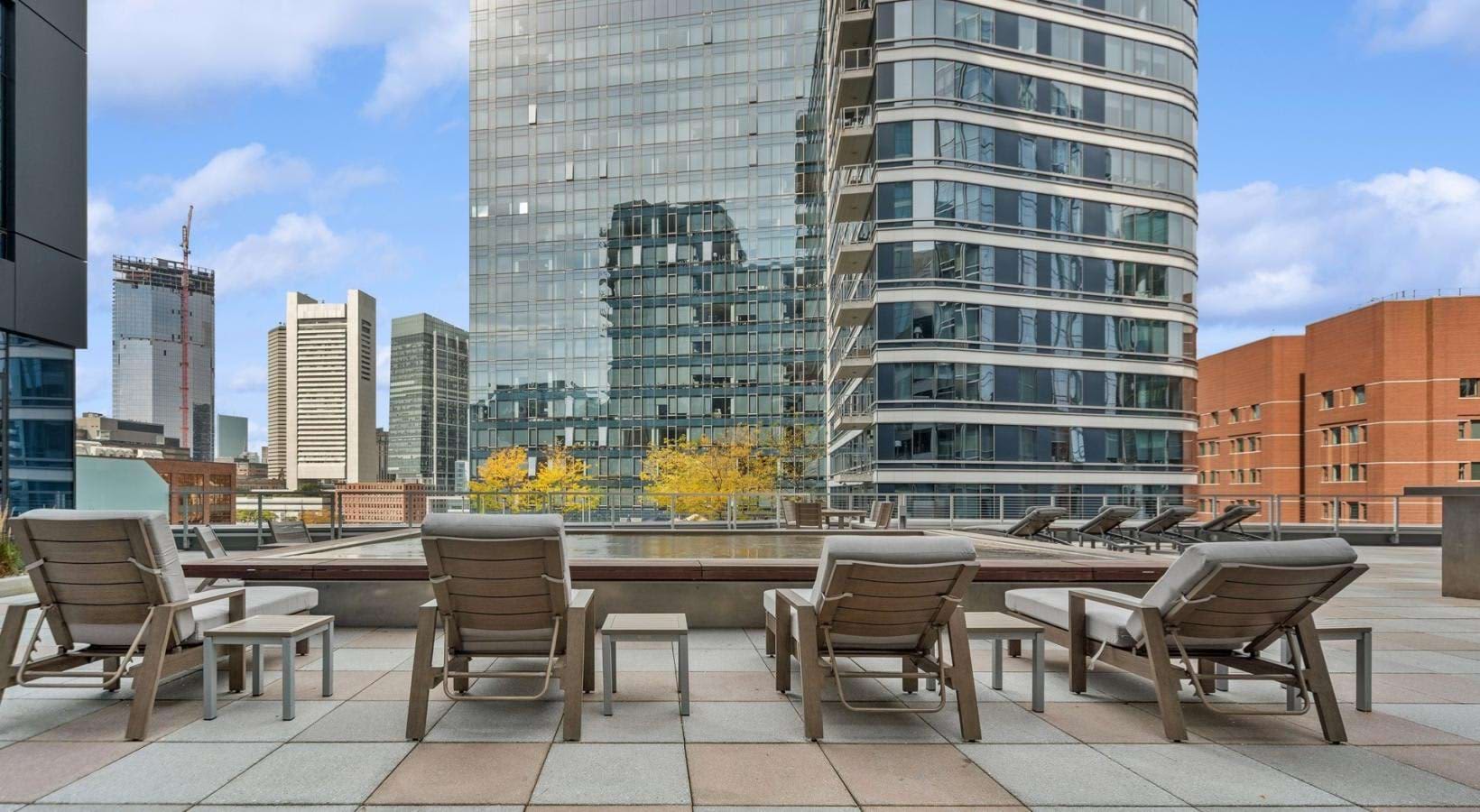 Rooftop terrace with four tan lounge chairs facing downtown Boston skyline and river, modern high-rise buildings visible.