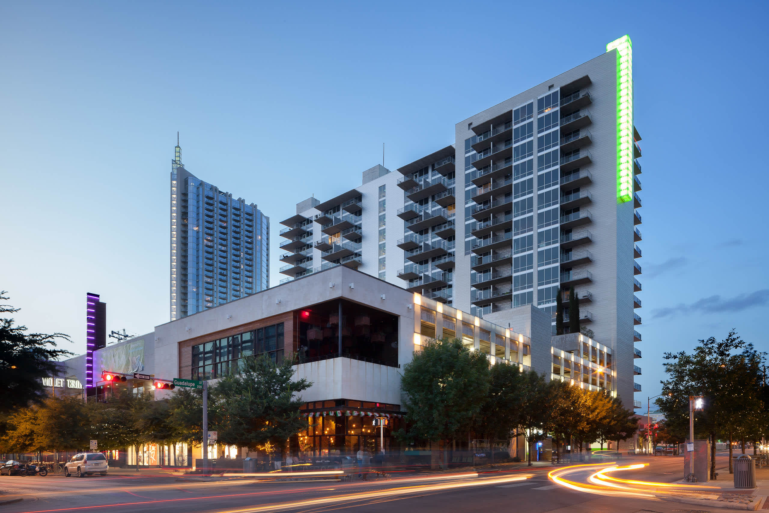 Modern multi-story residential building with ground-floor retail, illuminated storefront, and green accent lighting at dusk in urban setting.