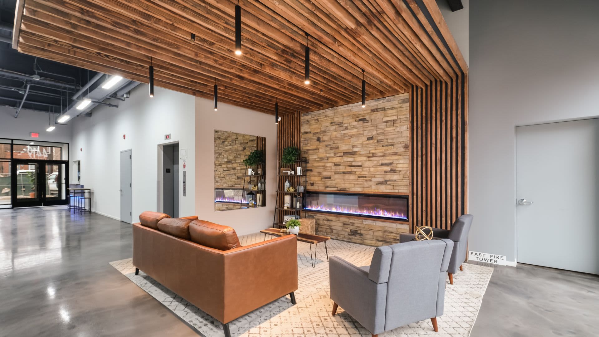 Common lounge with orange leather sofa, gray armchair, brick fireplace accent wall, and wood-slat ceiling with pendant lights.