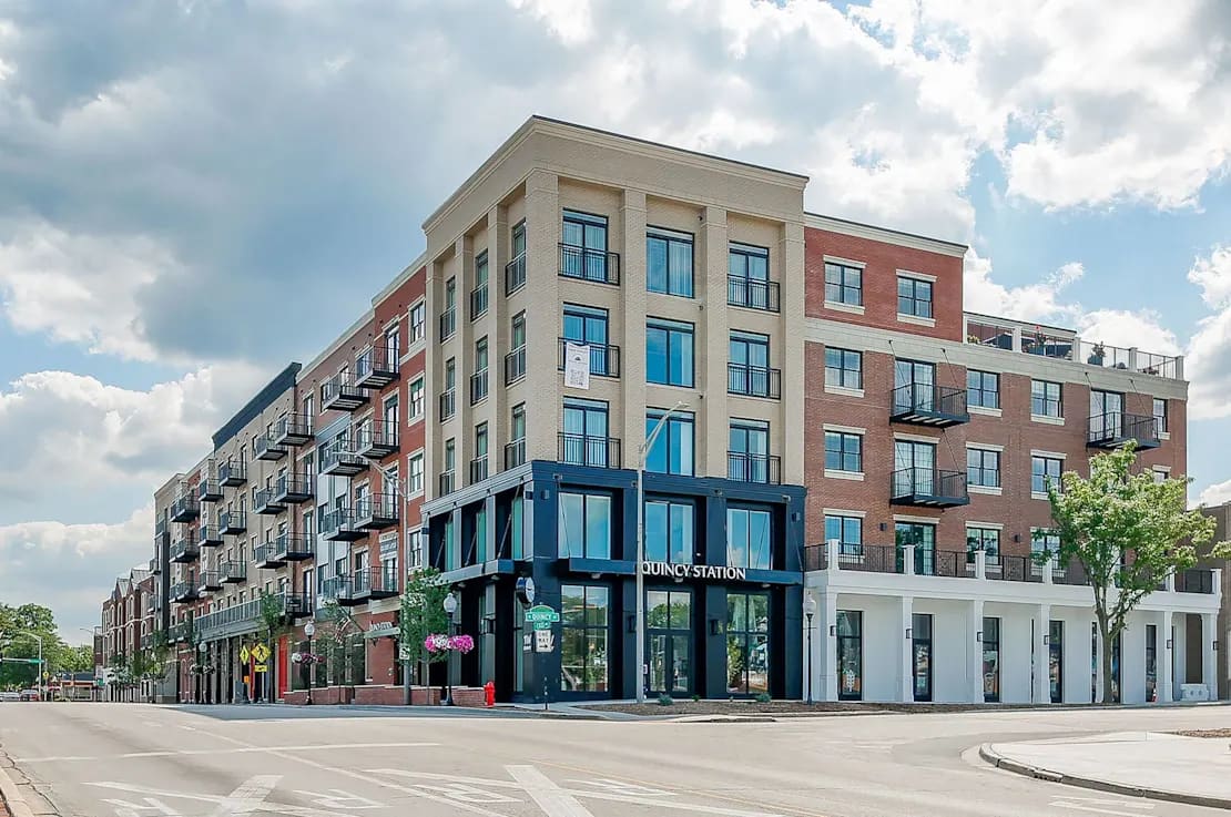 Multi-story residential building with brick and tan facade, ground-floor retail storefront labeled Quincy Station, black storefront windows, and metal balconies.