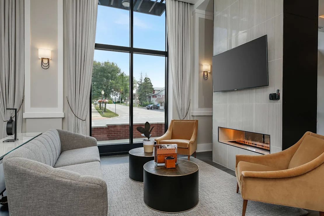 Living room with gray curved sofa, tan armchairs, black round tables, wall-mounted TV, gas fireplace, and large windows overlooking street.