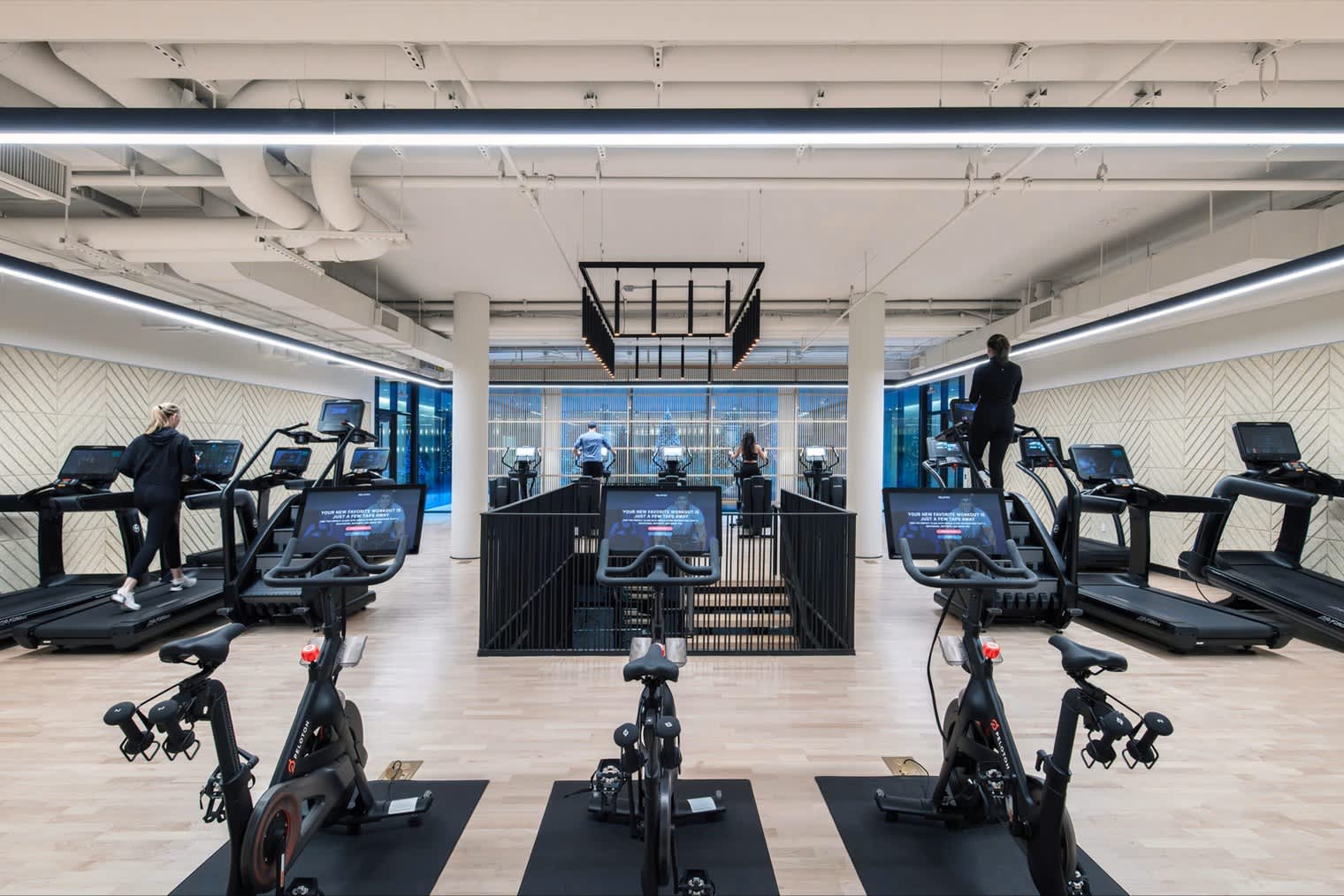Fitness center with stationary bikes in foreground, treadmills and cardio machines along sides, large windows overlooking city, exposed ceiling with linear lighting.