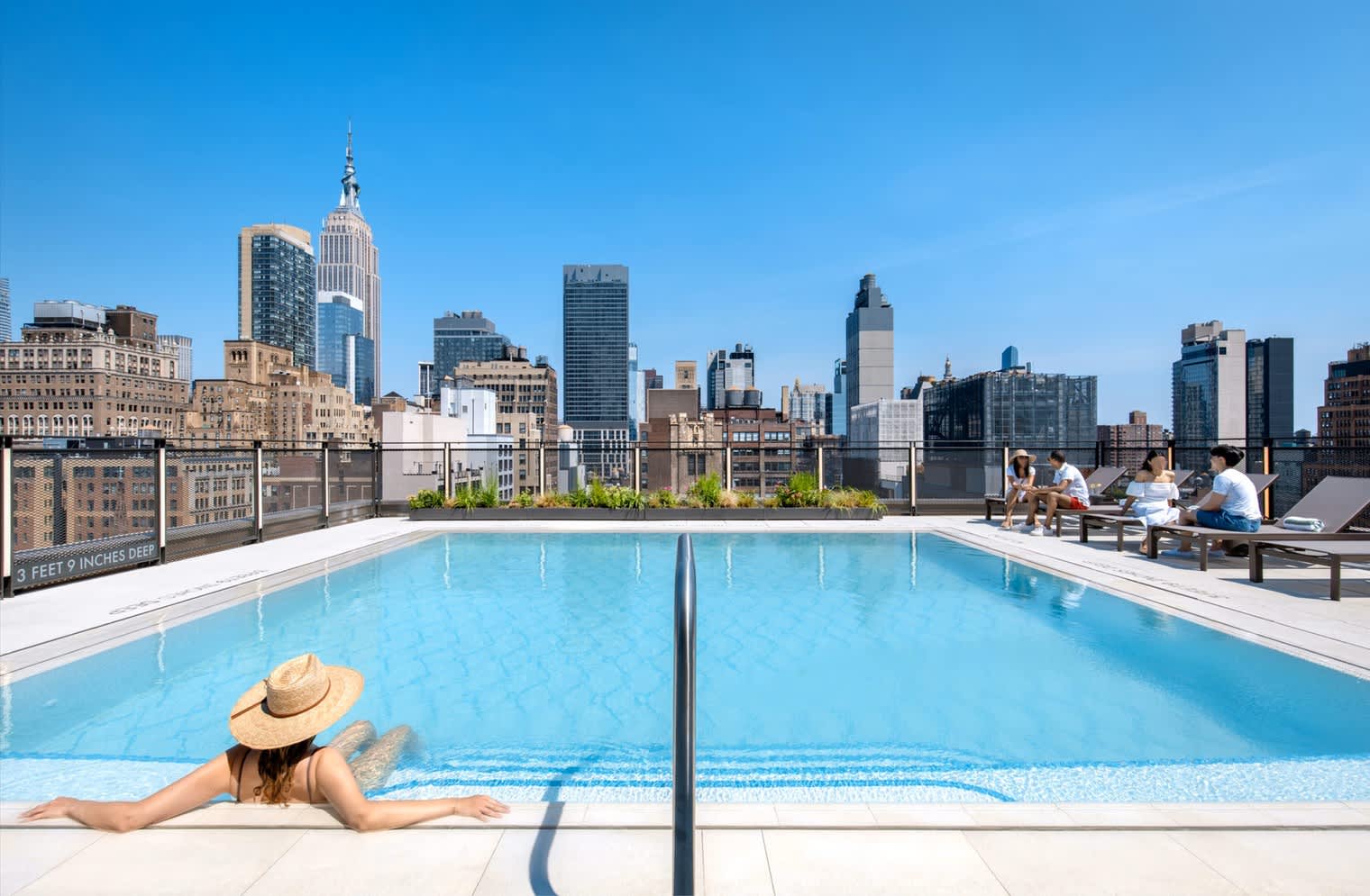Rooftop swimming pool with Manhattan skyline featuring Empire State Building, lounge seating, and residents enjoying the deck.