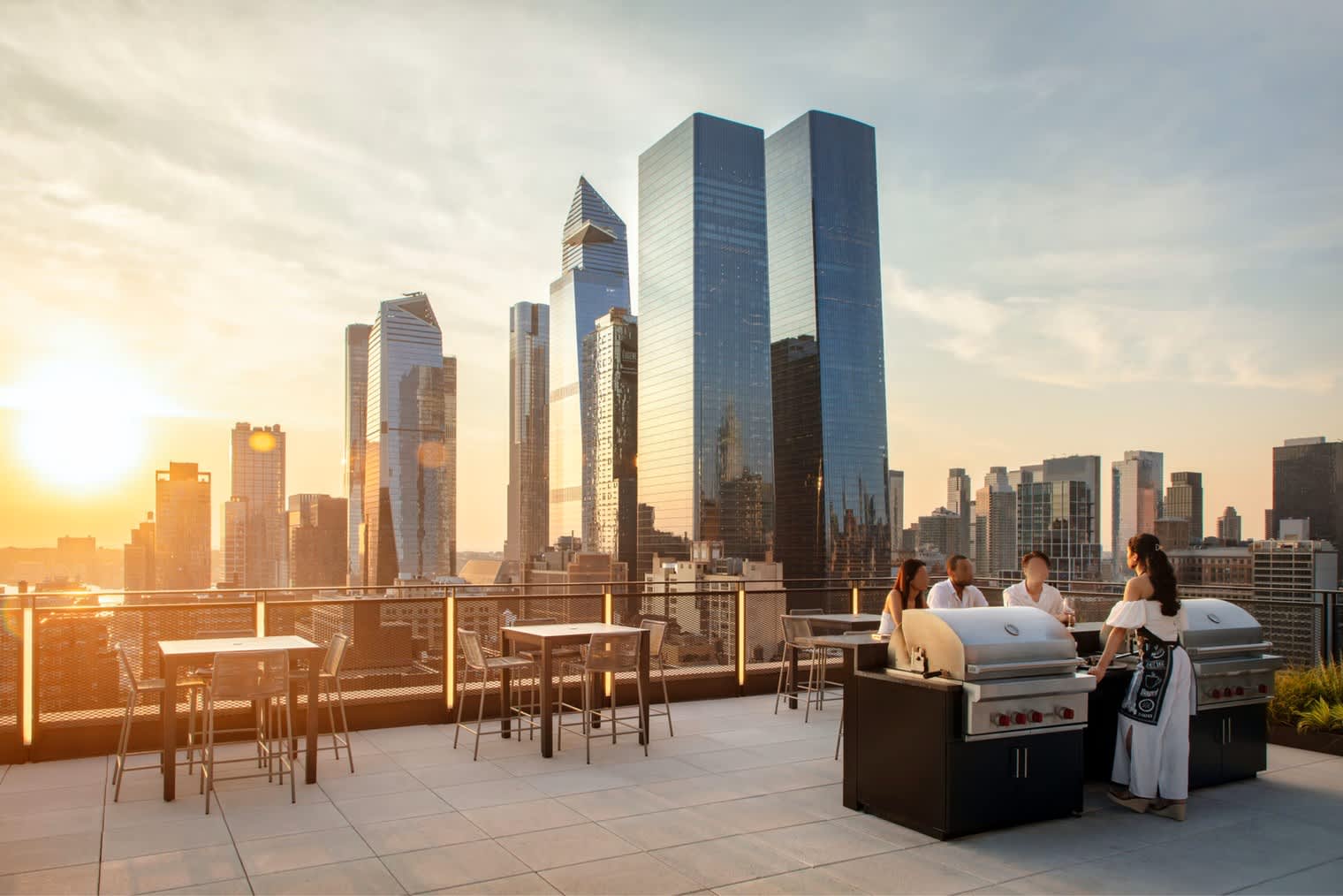 Rooftop terrace with dining tables, built-in grills, and people gathered overlooking downtown Manhattan skyscrapers at golden hour.