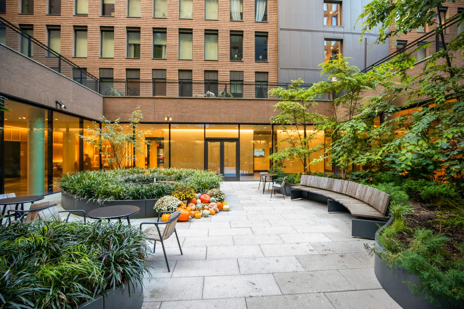 Brick courtyard with paved stone flooring, curved benches, potted plants, ivy-covered walls, and illuminated ground-floor windows of surrounding buildings.
