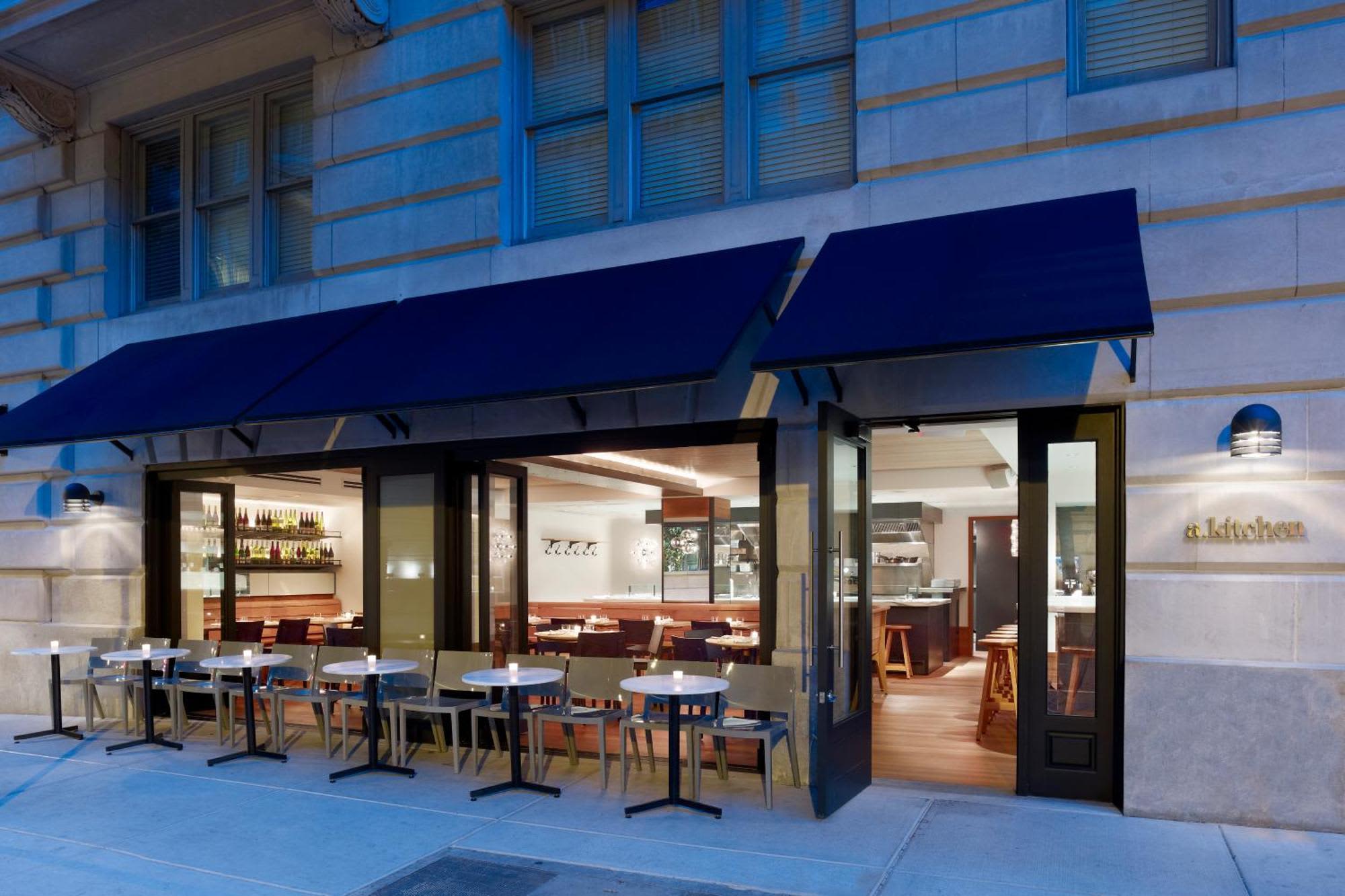 Restaurant exterior with blue awning, outdoor seating, and illuminated interior bar and dining area visible through large windows.