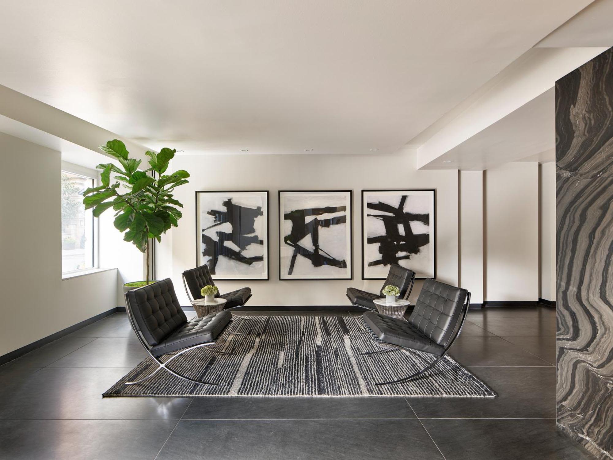 Modern lounge with two black leather chairs, striped area rug, potted green plant, and three framed abstract artworks on white walls.
