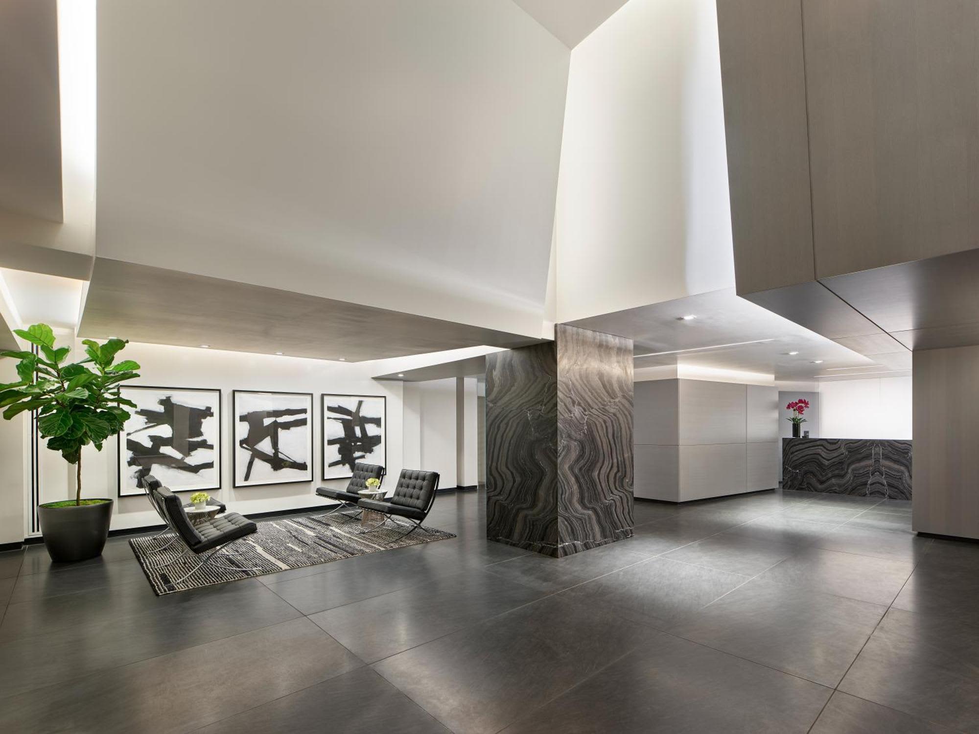 Modern lobby with polished concrete floors, sculptural marble column, contemporary seating, framed artwork, and potted plant under geometric ceiling design.