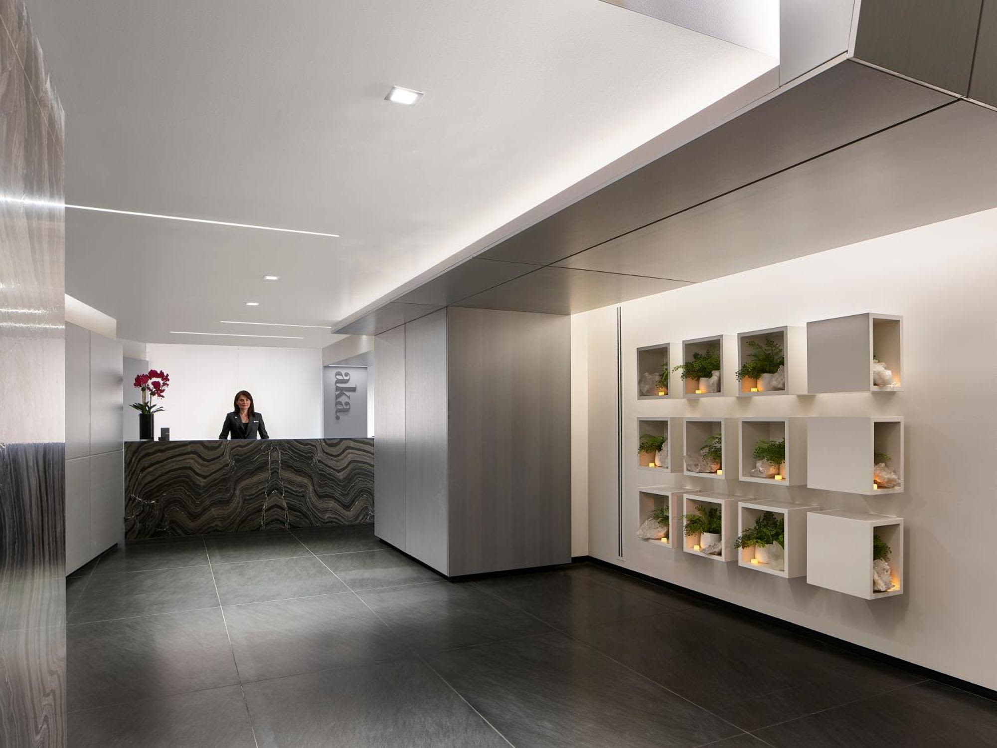 Modern lobby with curved black marble reception desk, staff member, white wall shelving with potted plants, and recessed ceiling lights.