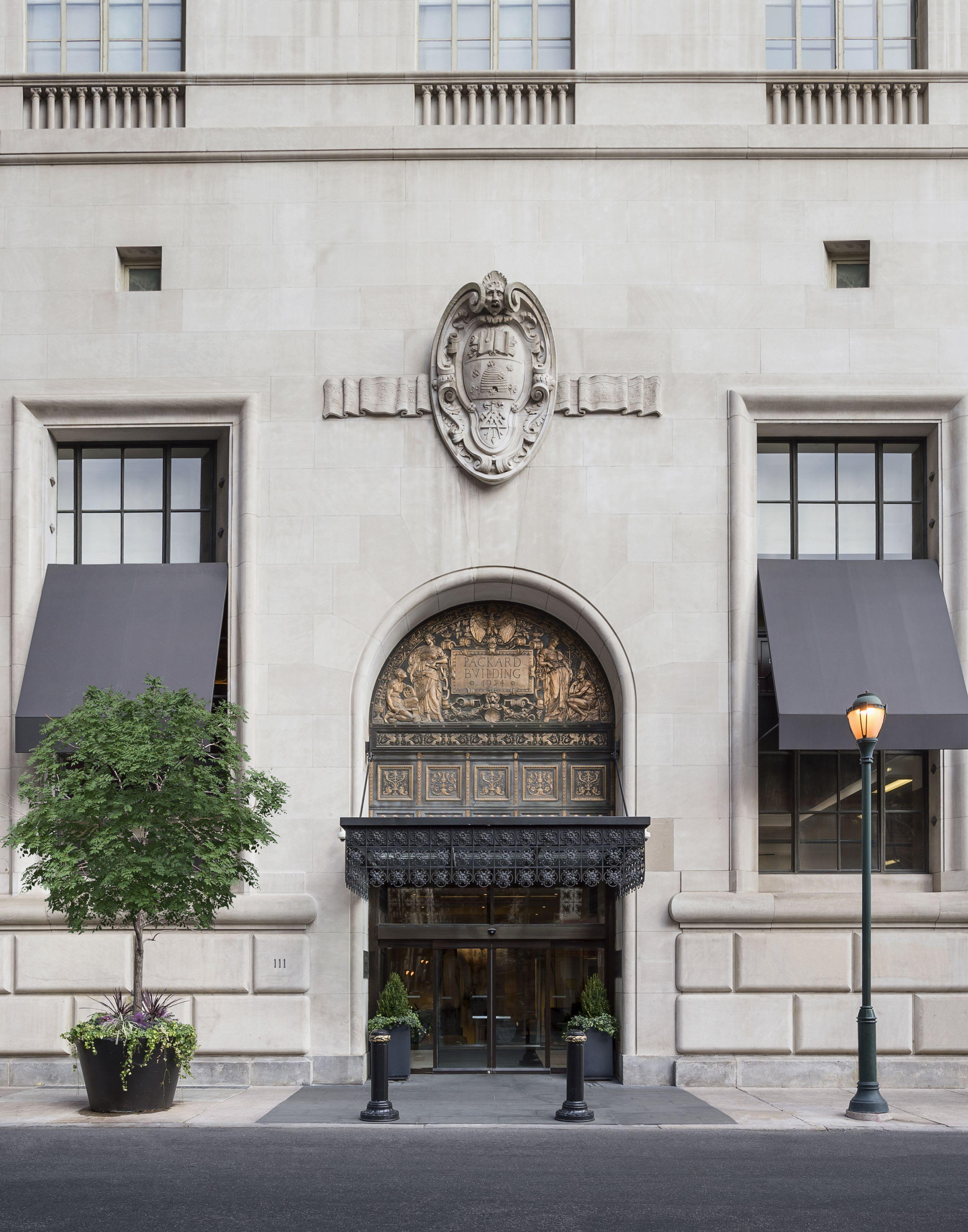 Historic limestone building facade with arched bronze doorway, ornamental crest, symmetrical windows with awnings, and planted greenery flanking the entrance.