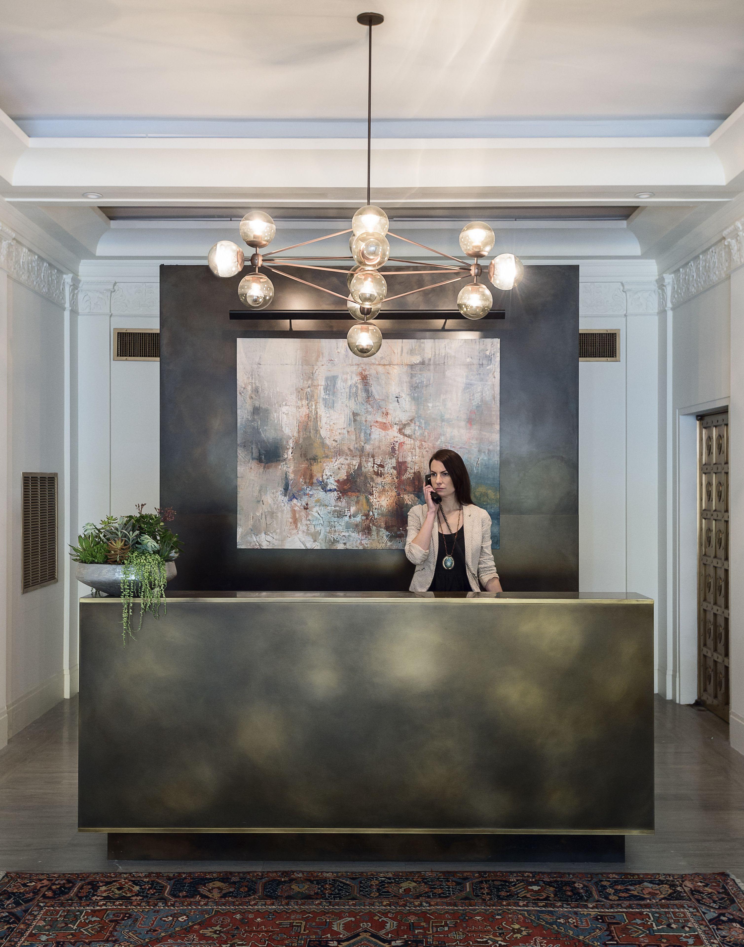 Lobby with dark reception desk, woman staff member, geometric brass pendant chandelier, abstract painting on black accent wall, potted plants, and coffered ceiling.
