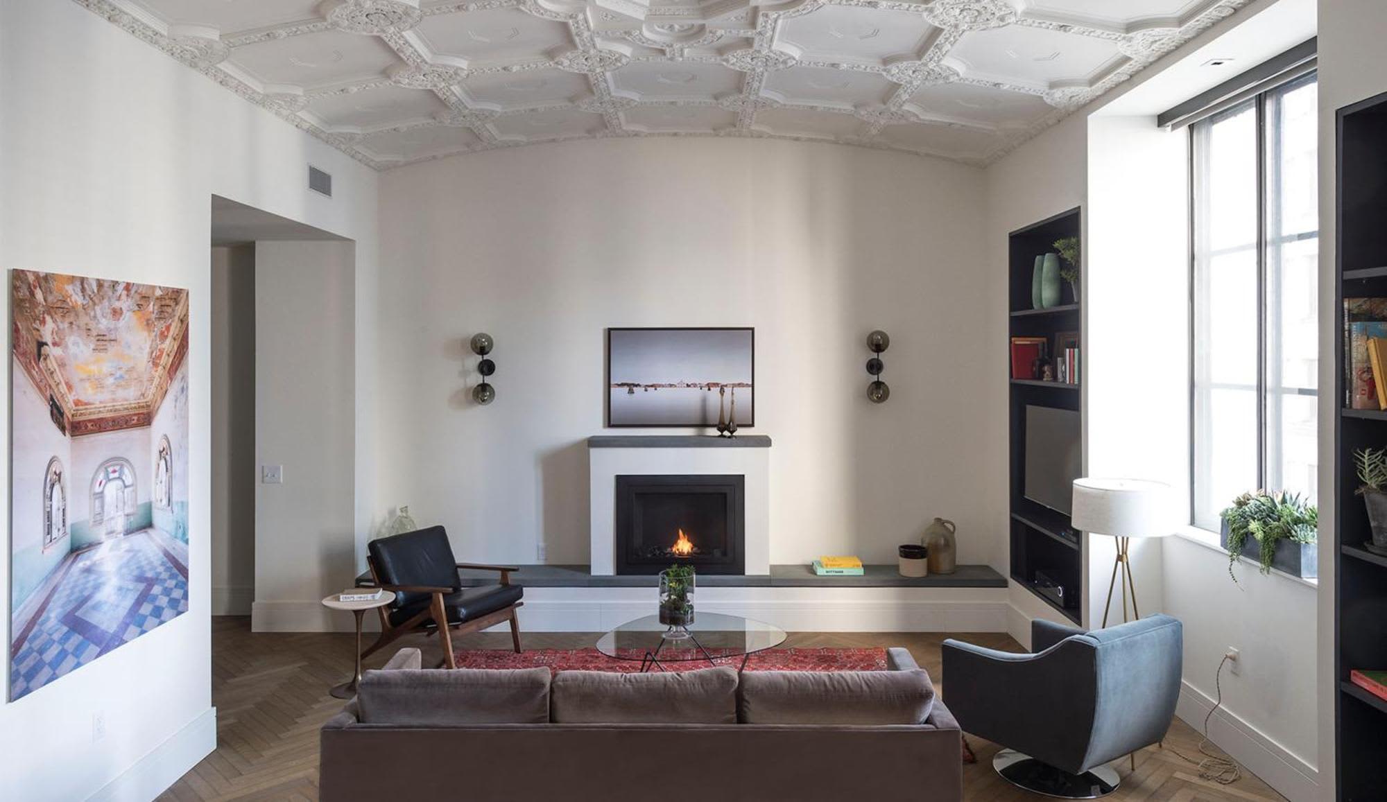 Modern living room with gray sofa, fireplace with white surround, black accent chairs, built-in shelving, and large windows with natural light.