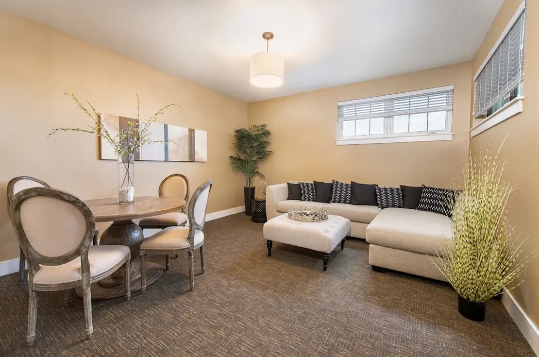 Open lounge with cream sectional sofa, round dining table with chairs, potted plants, and large windows with natural light.