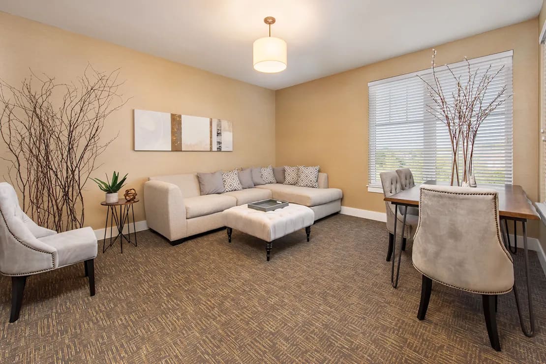 Spacious living room with beige sectional sofa, cream accent chairs, herringbone wood flooring, and large windows with plantation shutters.