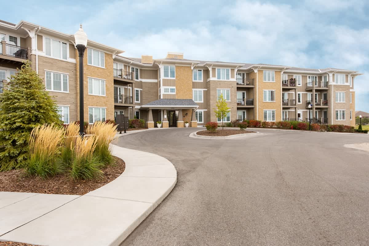 Multi-story brick apartment complex with curved driveway, landscaped entry with ornamental grasses, and covered building entrance.