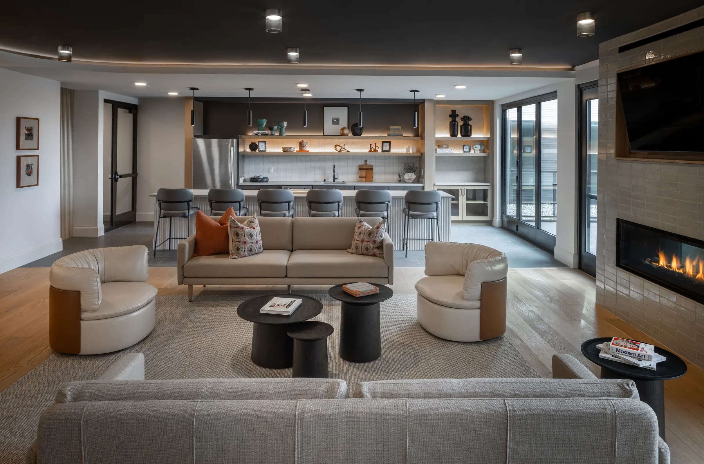 Open living room with beige sectional sofa, curved accent chairs, black side tables, and kitchen with white cabinetry and stainless steel appliances visible in background.
