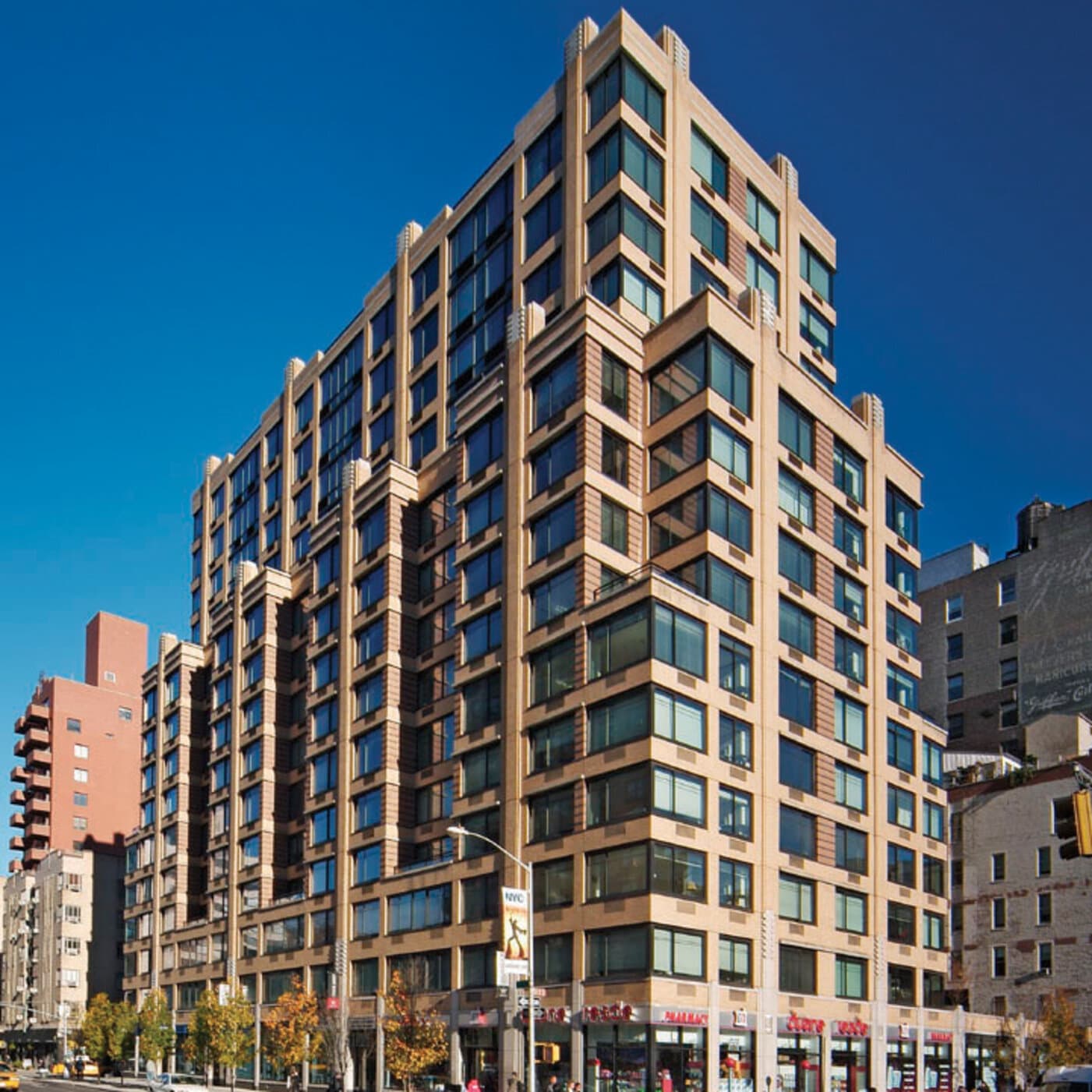 Tan brick residential building with regular window grid pattern, ground-floor retail storefronts, and neighboring buildings under clear blue sky.