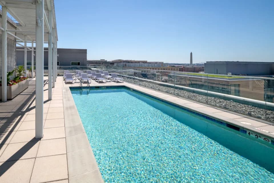 Rooftop pool with clear blue water, white lounge chairs, pergola shade structure, and Washington Monument visible in the distance.