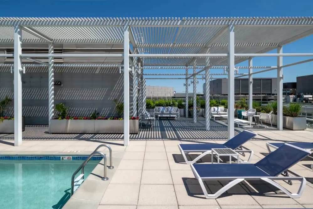 Rooftop pool area with white pergolas, lounge chairs, planters, and clear blue sky overlooking the city.