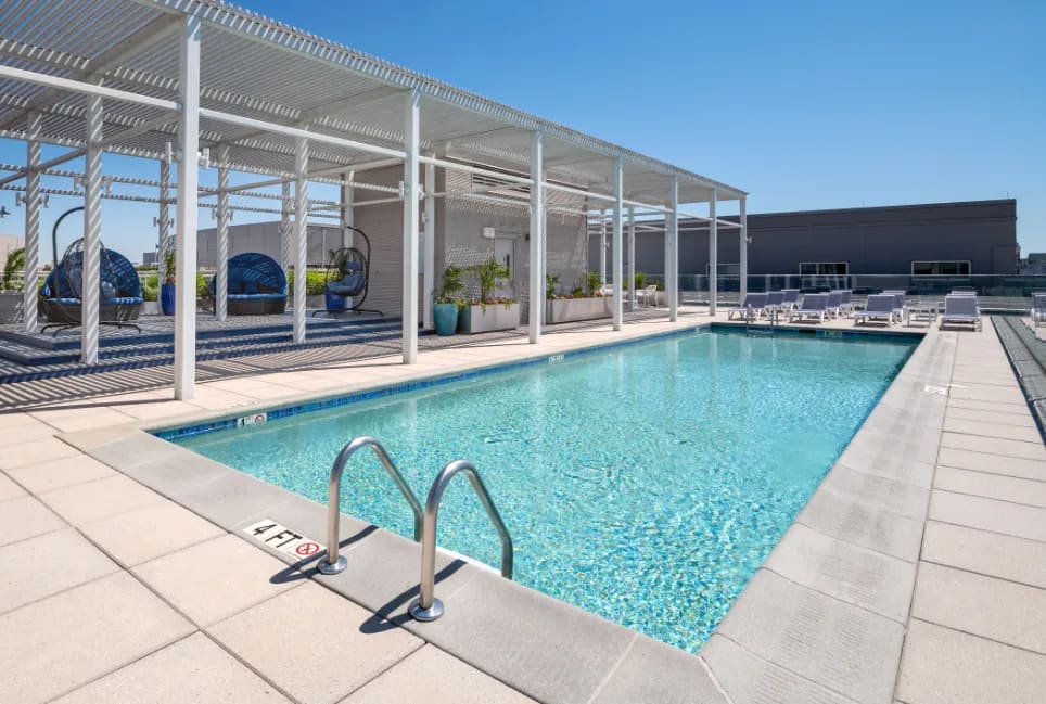 Rooftop pool with clear turquoise water, metal ladder in foreground, white pergola structure with lounge chairs and potted plants, city skyline visible in background.