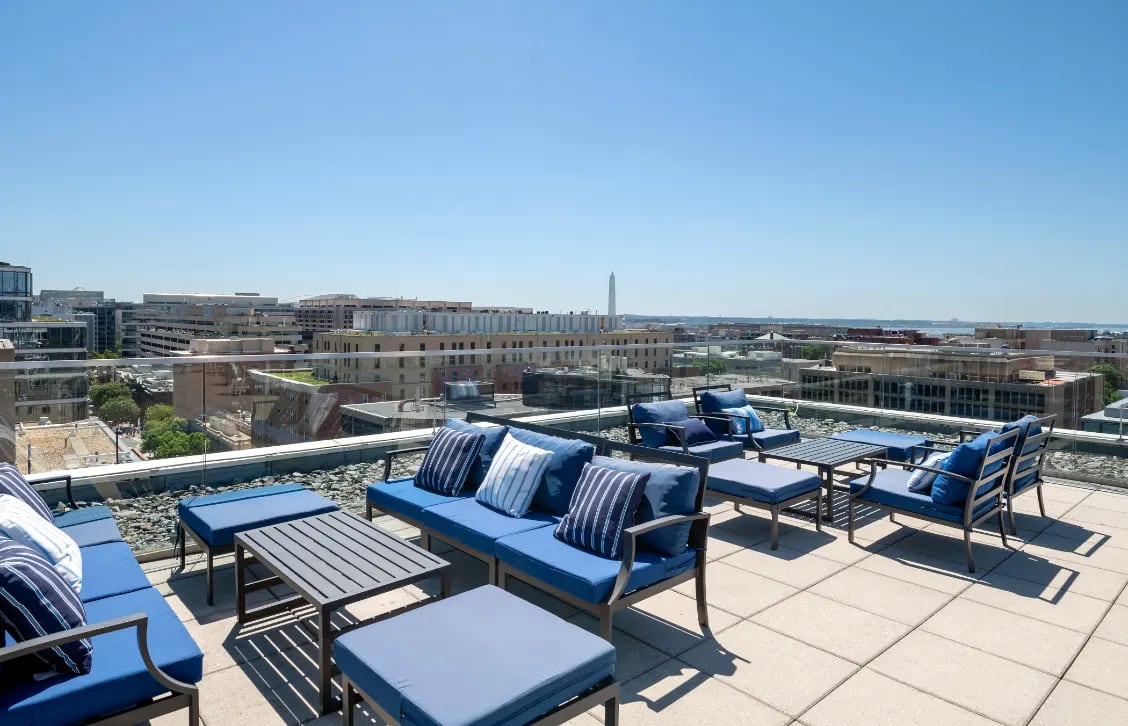 Rooftop lounge with blue cushioned seating, striped pillows, and metal tables overlooking Washington DC skyline and Washington Monument.