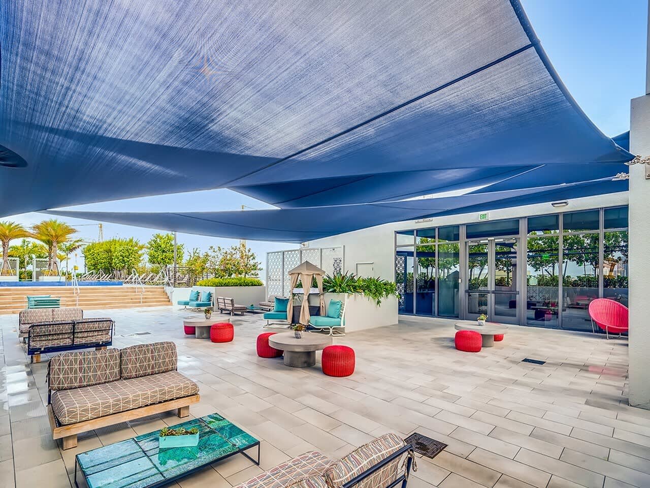 Rooftop terrace with blue shade sails, patterned seating, red poufs, and glass doors opening to interior spaces.
