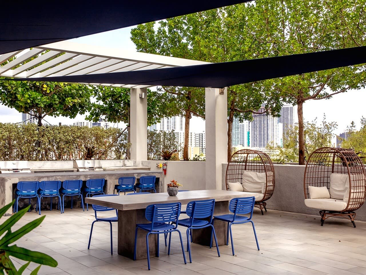 Rooftop terrace with blue dining chairs around a concrete table, two woven hanging chairs with cushions, retractable shade canopy, mature trees, and downtown Miami skyline in background.