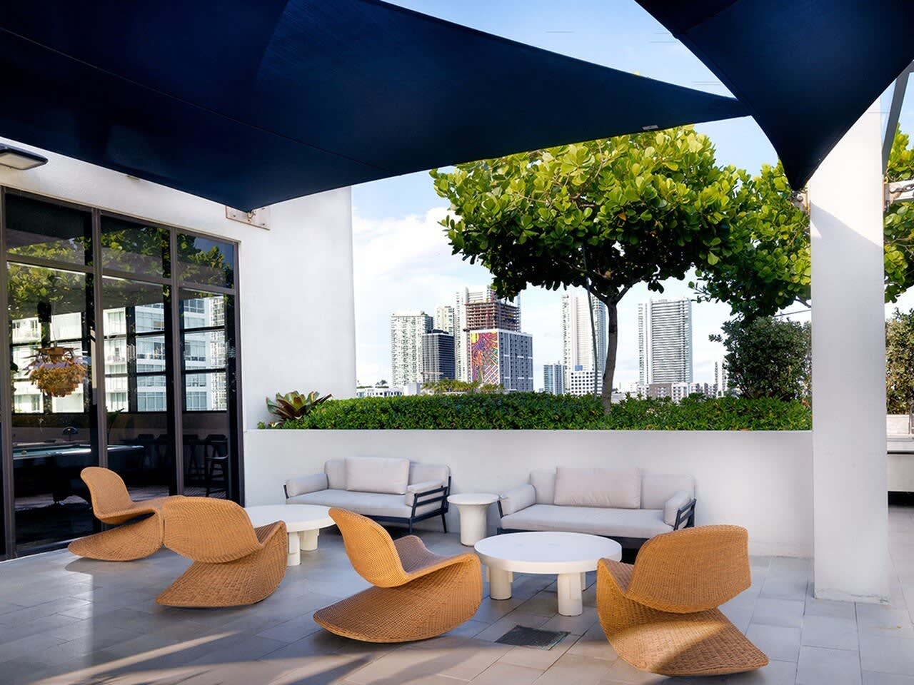 Rooftop terrace with woven orange lounge chairs, gray sectional sofa, and Miami skyline views beyond white walls and shade structure.
