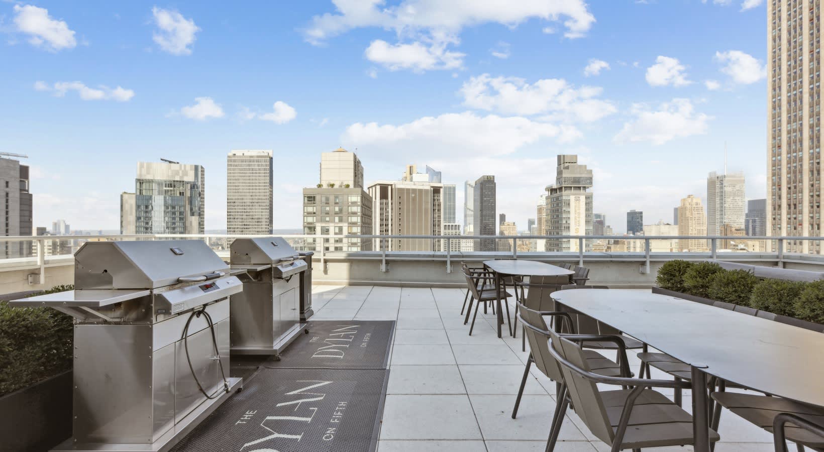 Rooftop terrace with built-in grills, dining tables, and chairs overlooking a dense urban skyline under blue sky.