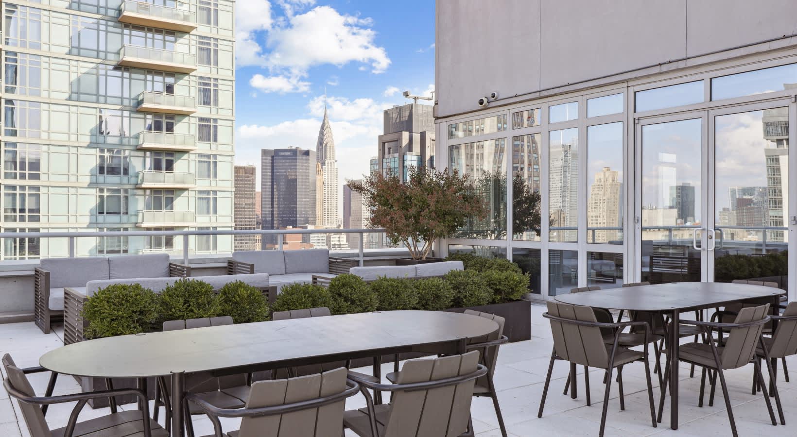 Rooftop terrace with dining tables, chairs, planters, and floor-to-ceiling windows overlooking Manhattan skyline and Chrysler Building.