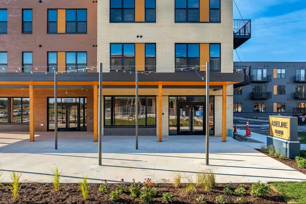 Modern apartment building exterior with orange and wood accents, ground-floor retail storefronts, and landscaped entry area.