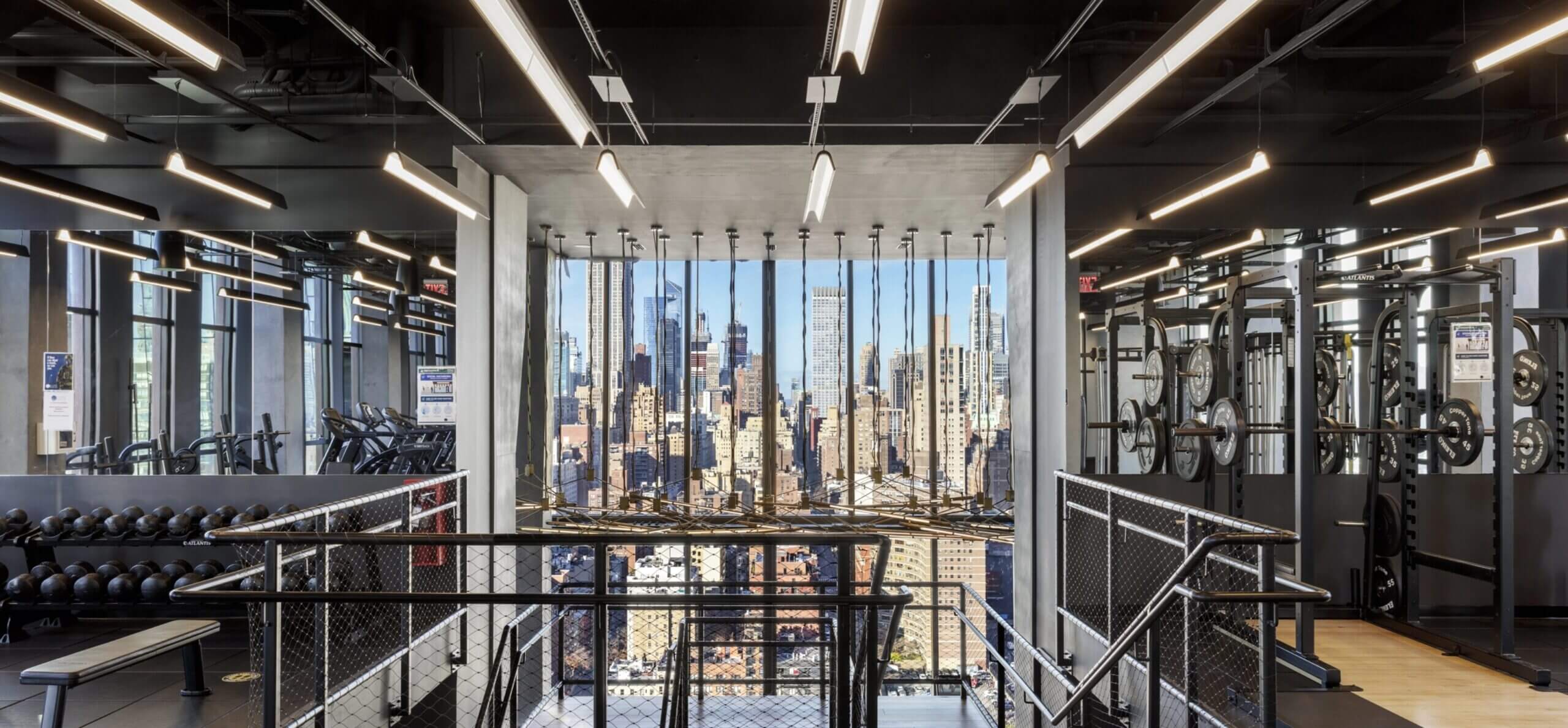 Modern gym with floor-to-ceiling windows framing Manhattan skyline, dumbbells on left, cardio machines center, bikes on right, industrial ceiling lights.