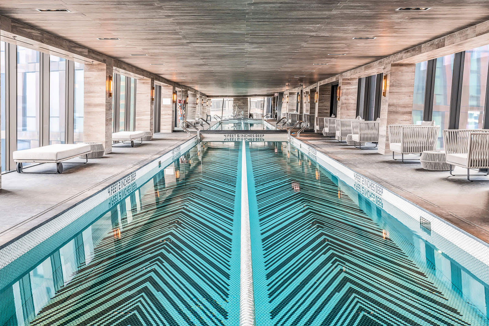 Indoor lap pool with turquoise and white geometric tile pattern, white lounge chairs, and floor-to-ceiling windows on both sides.