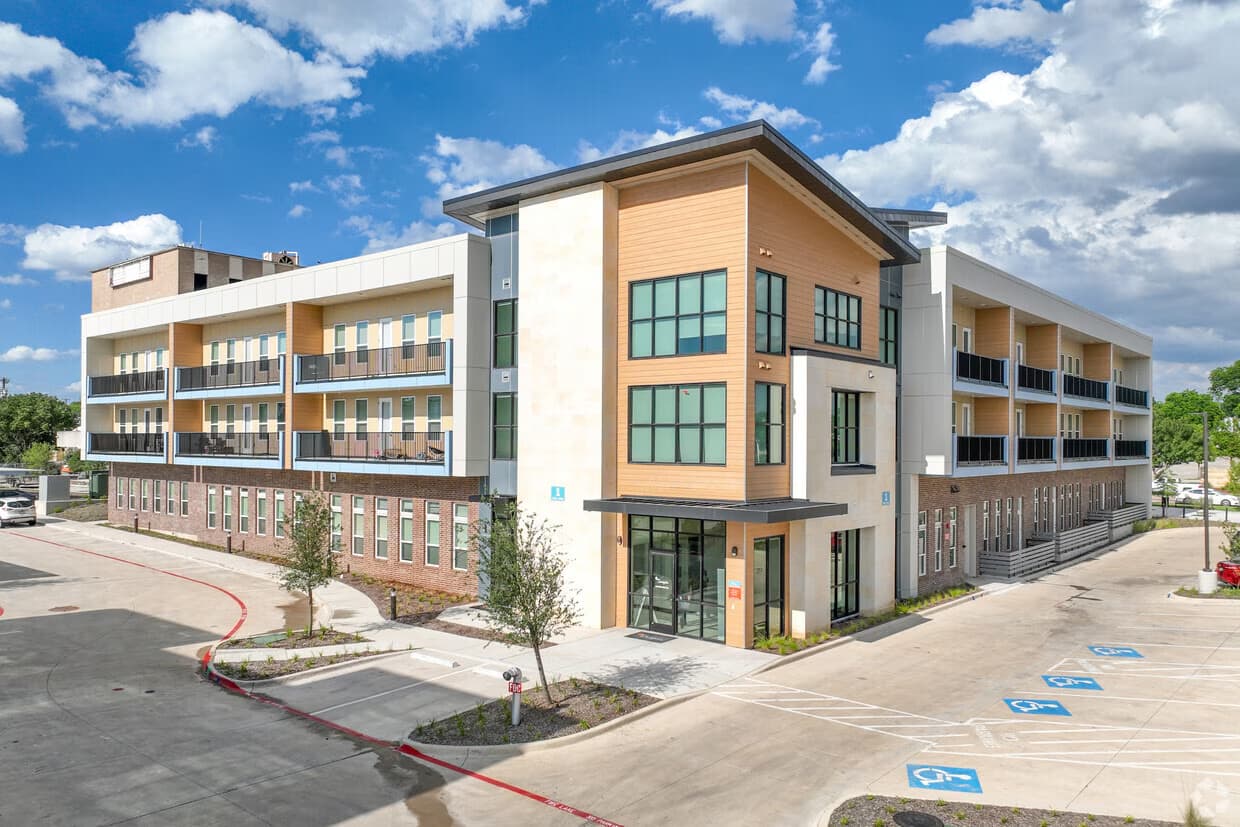 Four-story contemporary apartment complex with wood and white facade, large windows, ground-level commercial space, and surface parking lot.