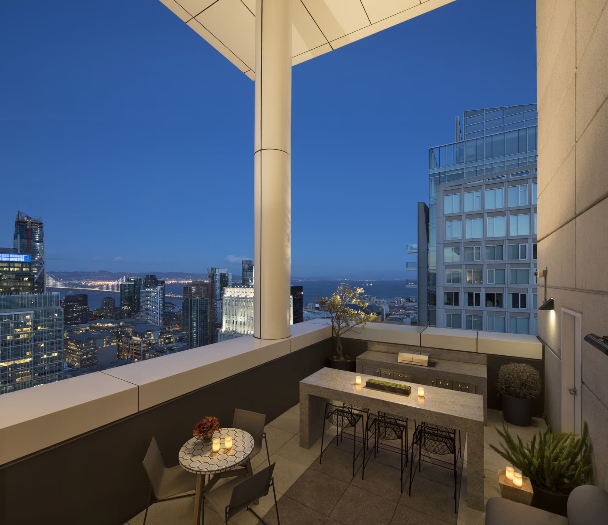 High-rise terrace with floor-to-ceiling windows overlooking San Francisco Bay, Golden Gate Bridge, city skyline, modern bar seating, and potted plants at dusk.