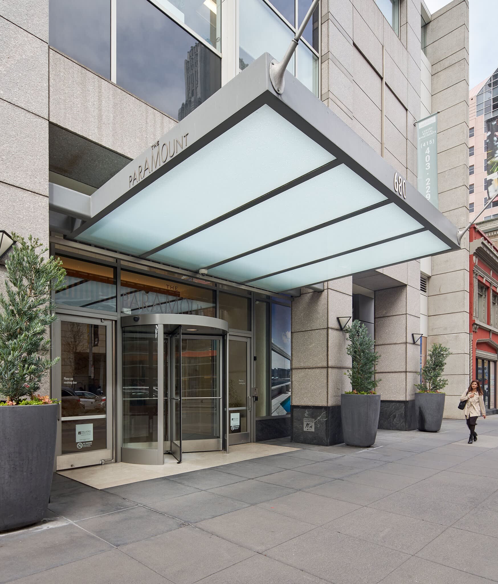 Building entrance with illuminated glass canopy, dark metal framing, glass doors, and potted plants flanking the entry.
