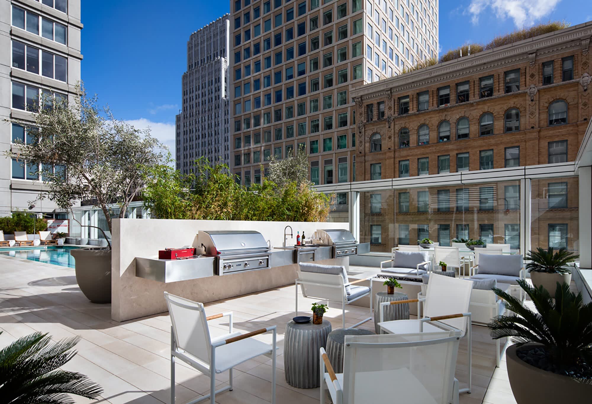 Rooftop terrace with outdoor kitchen, built-in grill, gray lounge chairs, potted plants, and San Francisco skyline backdrop.