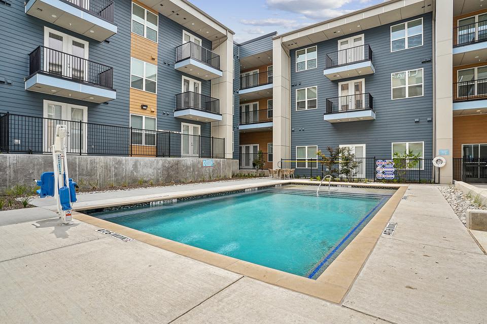 Rectangular swimming pool with turquoise water surrounded by concrete deck, flanked by multi-story apartment buildings with blue siding and metal balconies.