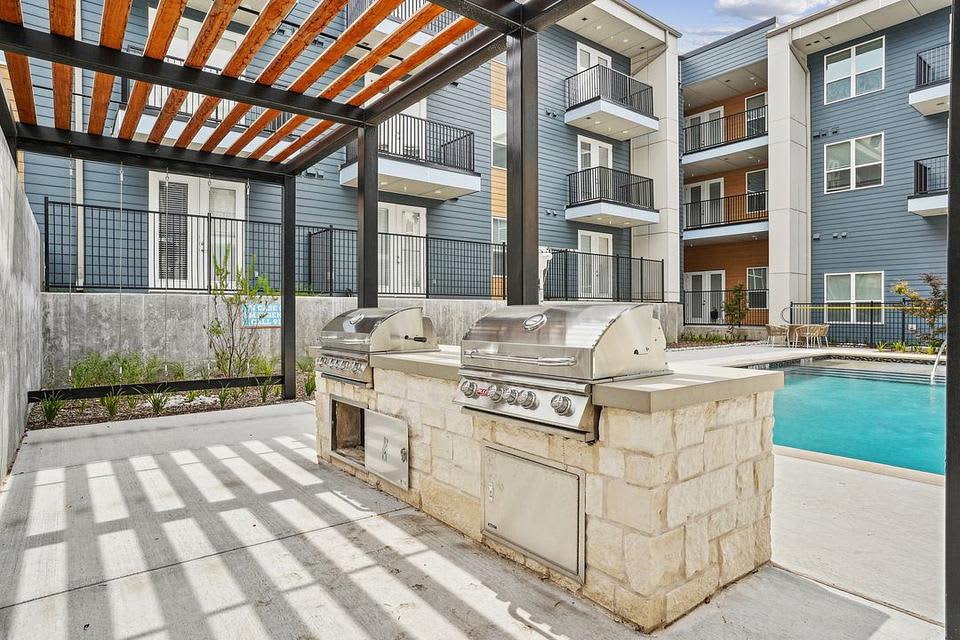 Covered patio with wooden pergola, stainless steel built-in grills on stone counter, and view of turquoise pool with multi-story residential buildings beyond.