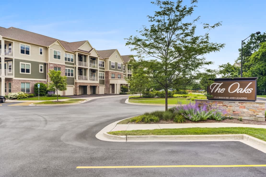 Multi-story apartment buildings with brick and siding exterior, landscaped entrance with purple flowers, and The Oaks community sign.