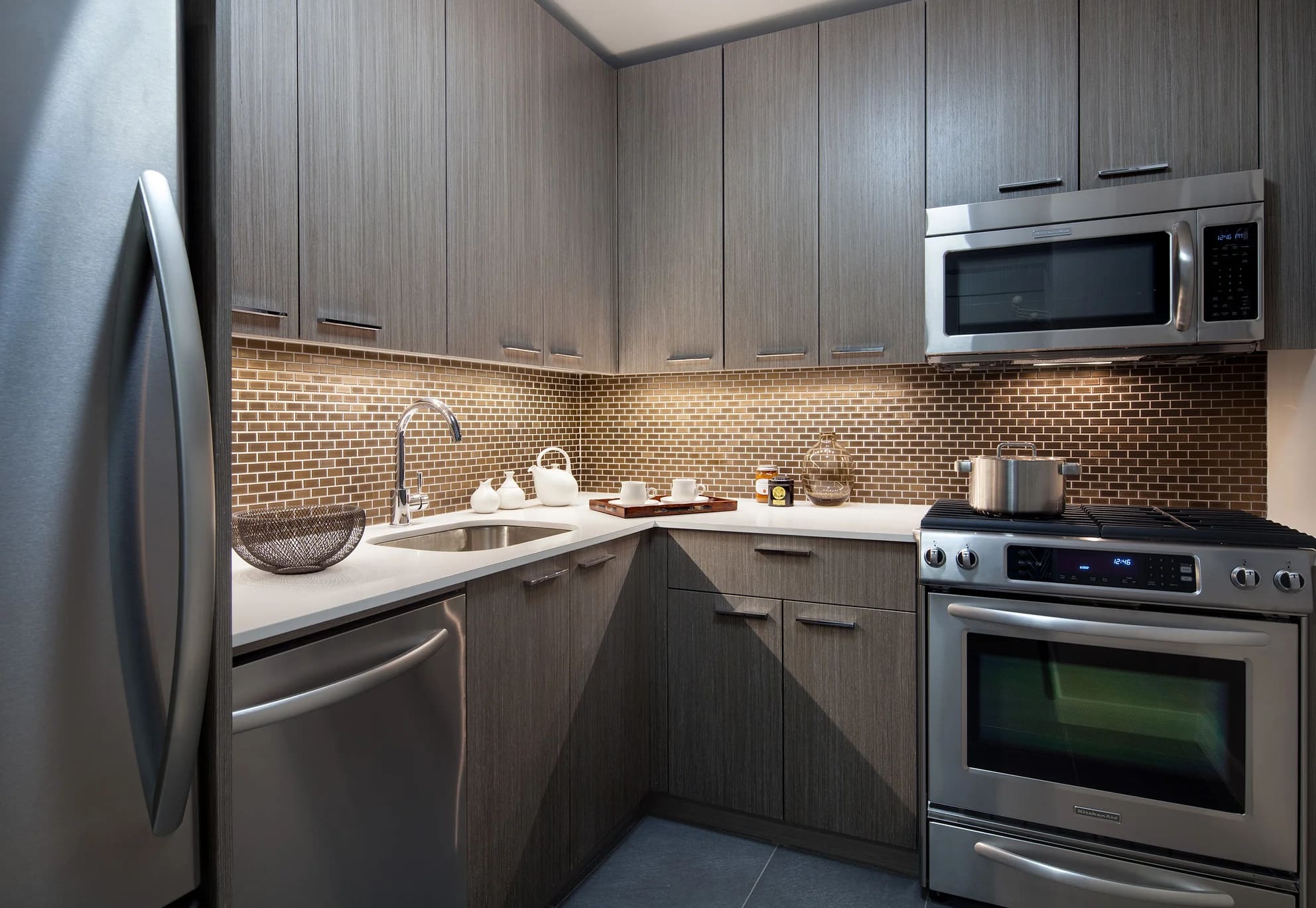 Modern kitchen with gray wood cabinets, stainless steel appliances, brick backsplash, and white countertops.