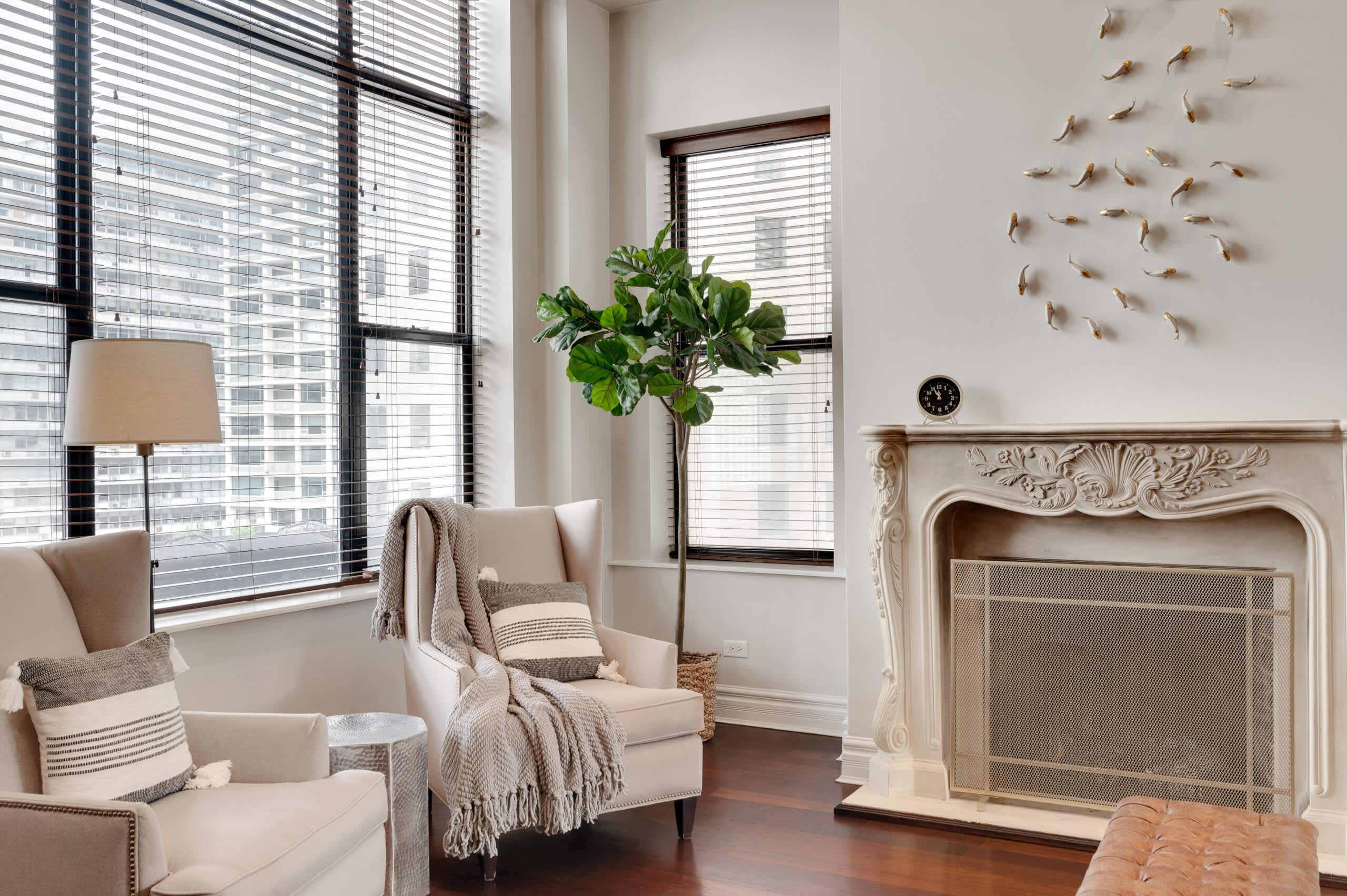 Living room with cream armchairs, decorative fireplace, potted plant, and floor-to-ceiling windows overlooking city buildings.