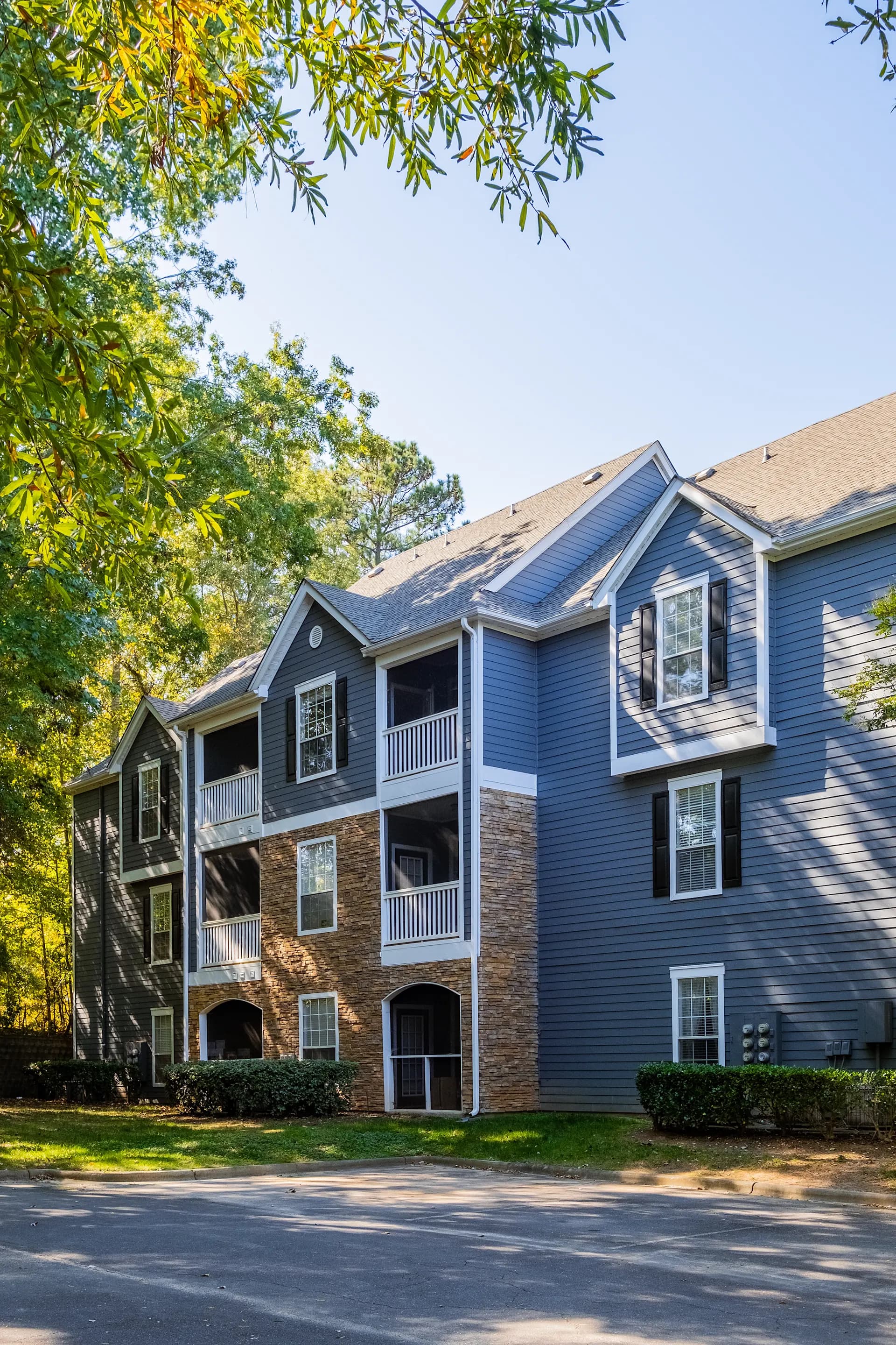 Three-story townhome with blue siding and brick base, white trim balconies, surrounded by mature trees and landscaping.