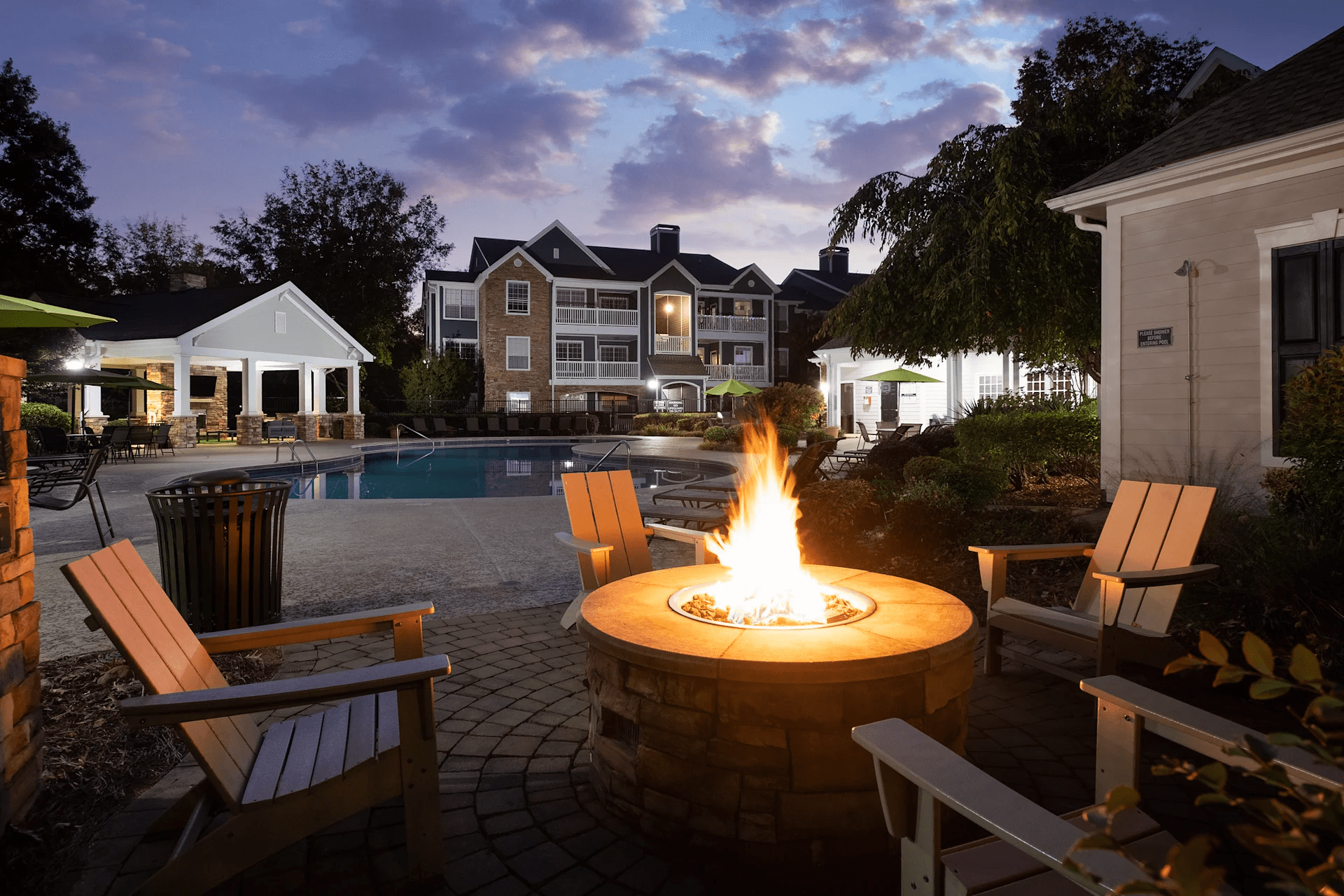 Outdoor fire pit with Adirondack chairs, illuminated pool, and residential buildings at dusk in Charlotte.